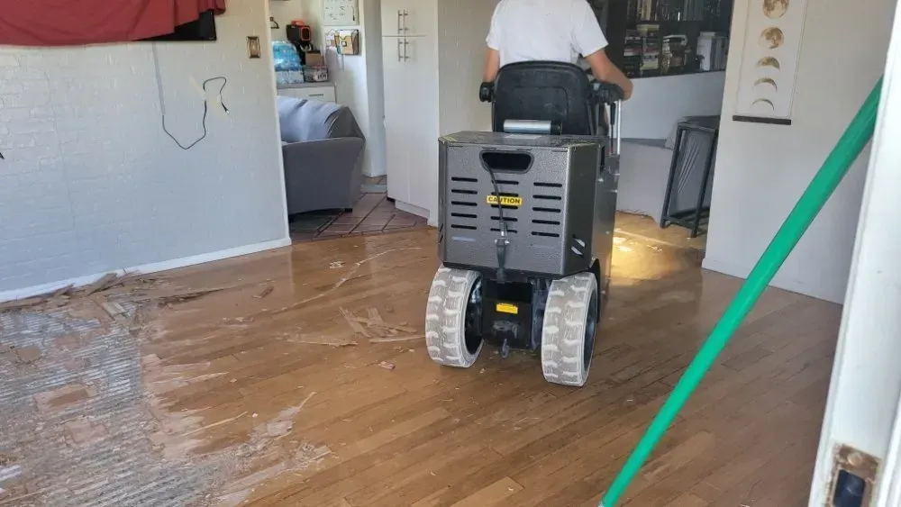 Person operating a floor scraping machine on a wet hardwood floor. A green hose is in view.