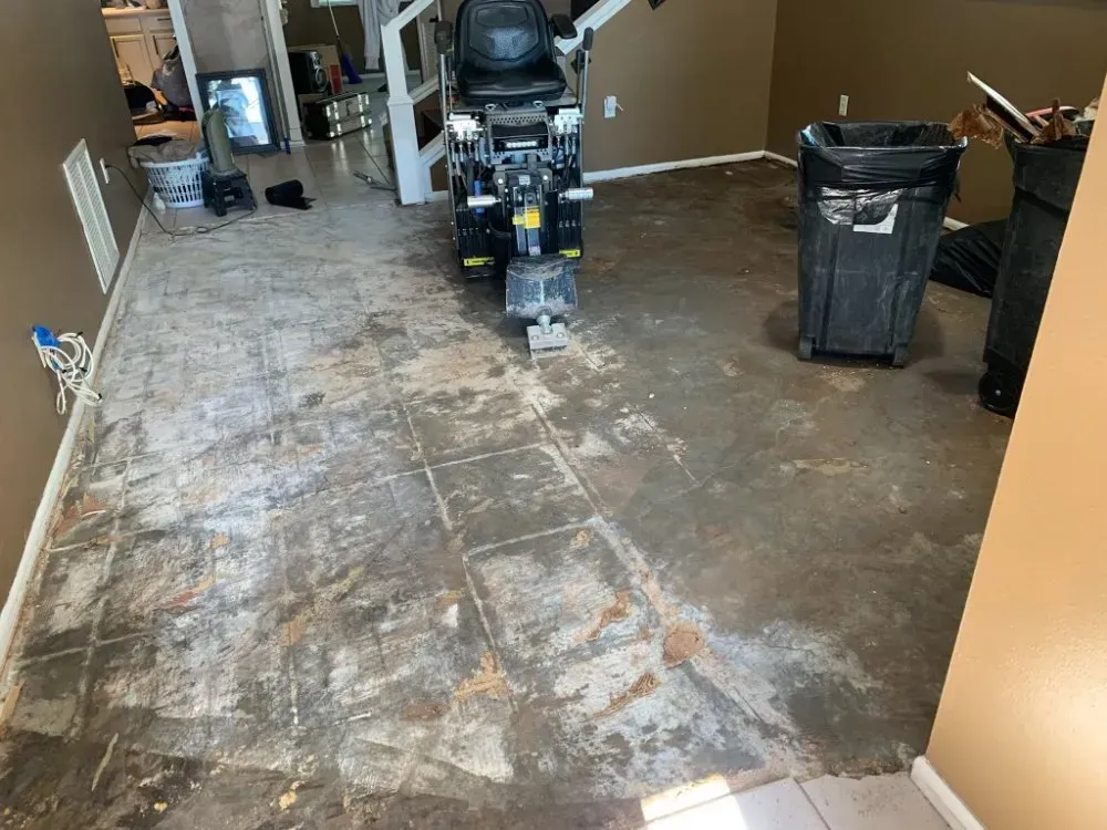 Room with floor tiles being removed by a machine. Brown walls, a trash can, and debris are visible.