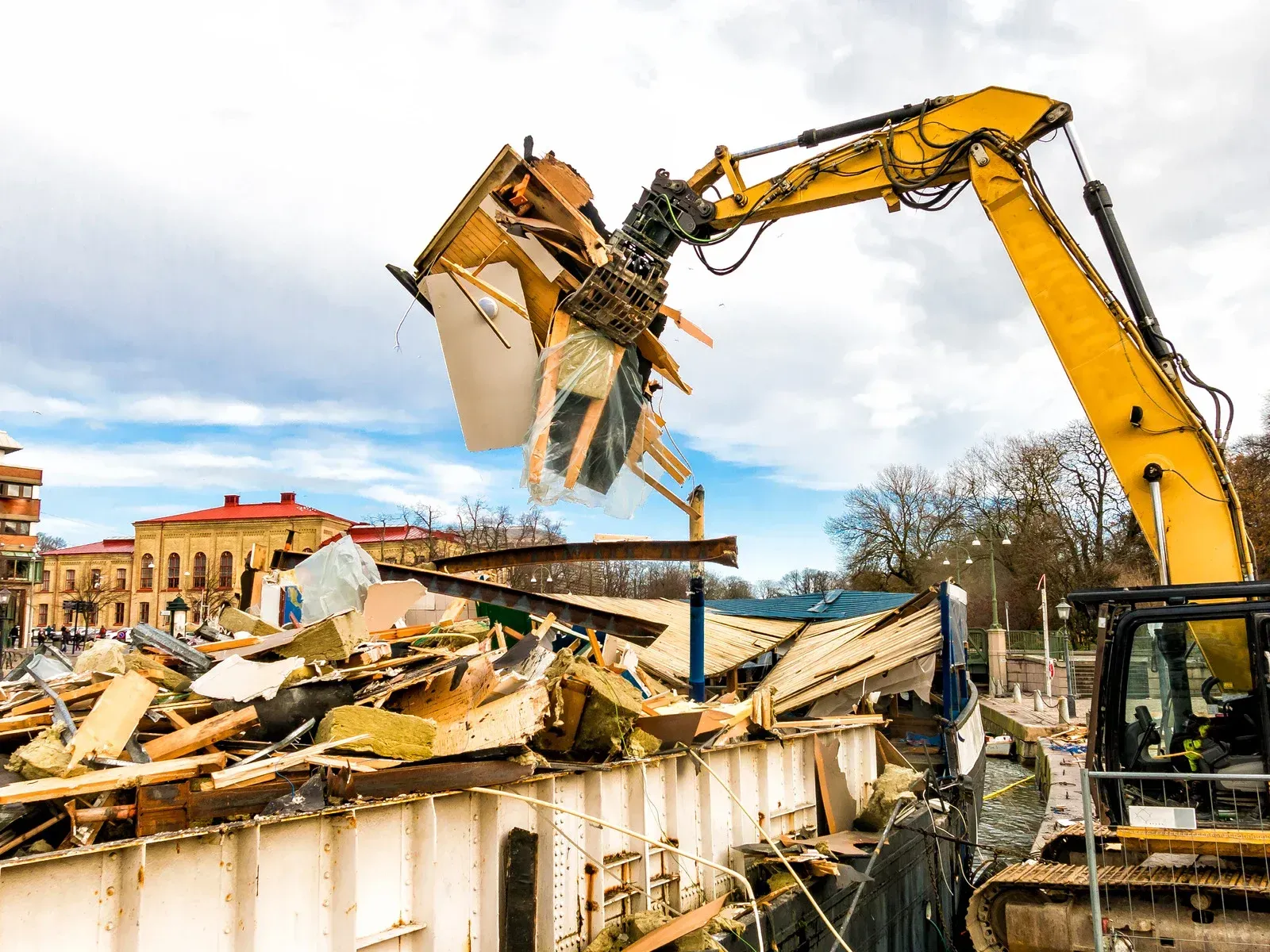 Yellow excavator demolishing a building with debris falling in a canal, cloudy sky.