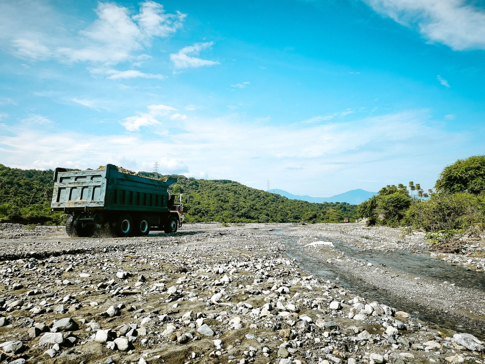 Dump truck driving on a rocky road, surrounded by greenery under a blue sky.