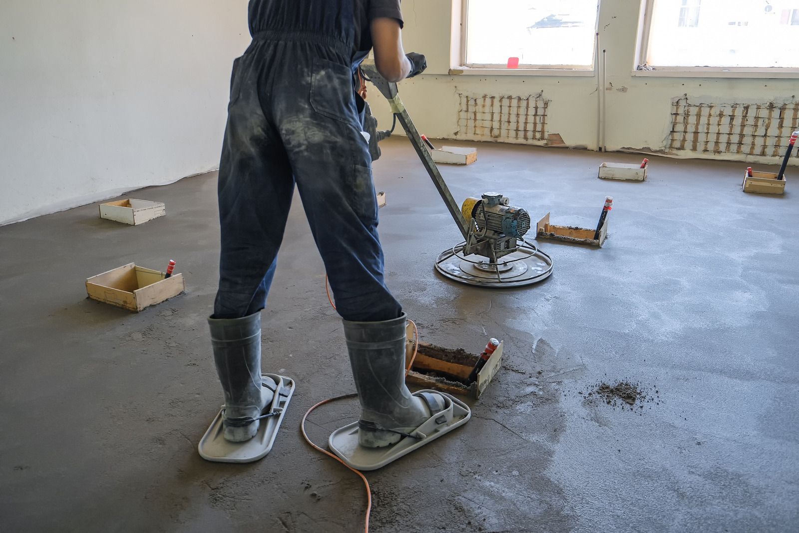 Person in overalls uses a power trowel on a concrete floor, standing on floatation stilts.