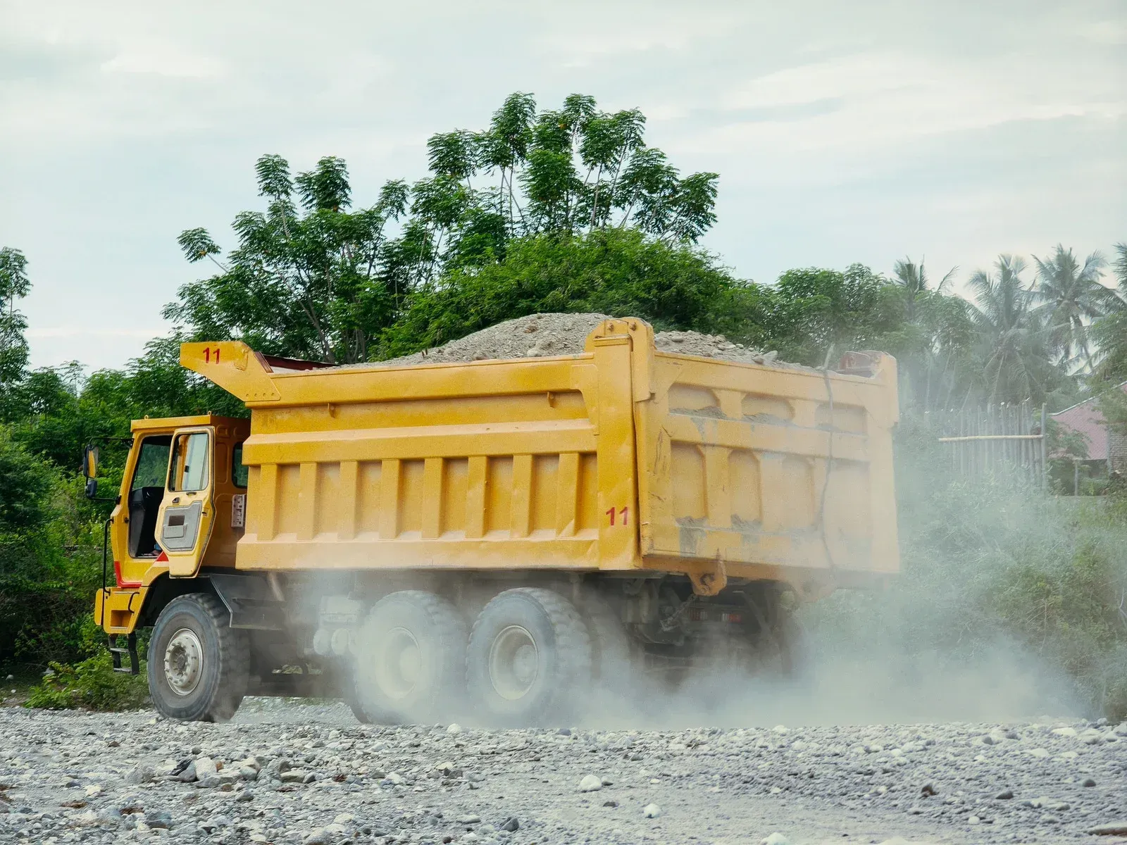 Yellow dump truck filled with gravel kicking up dust on a gravel road.