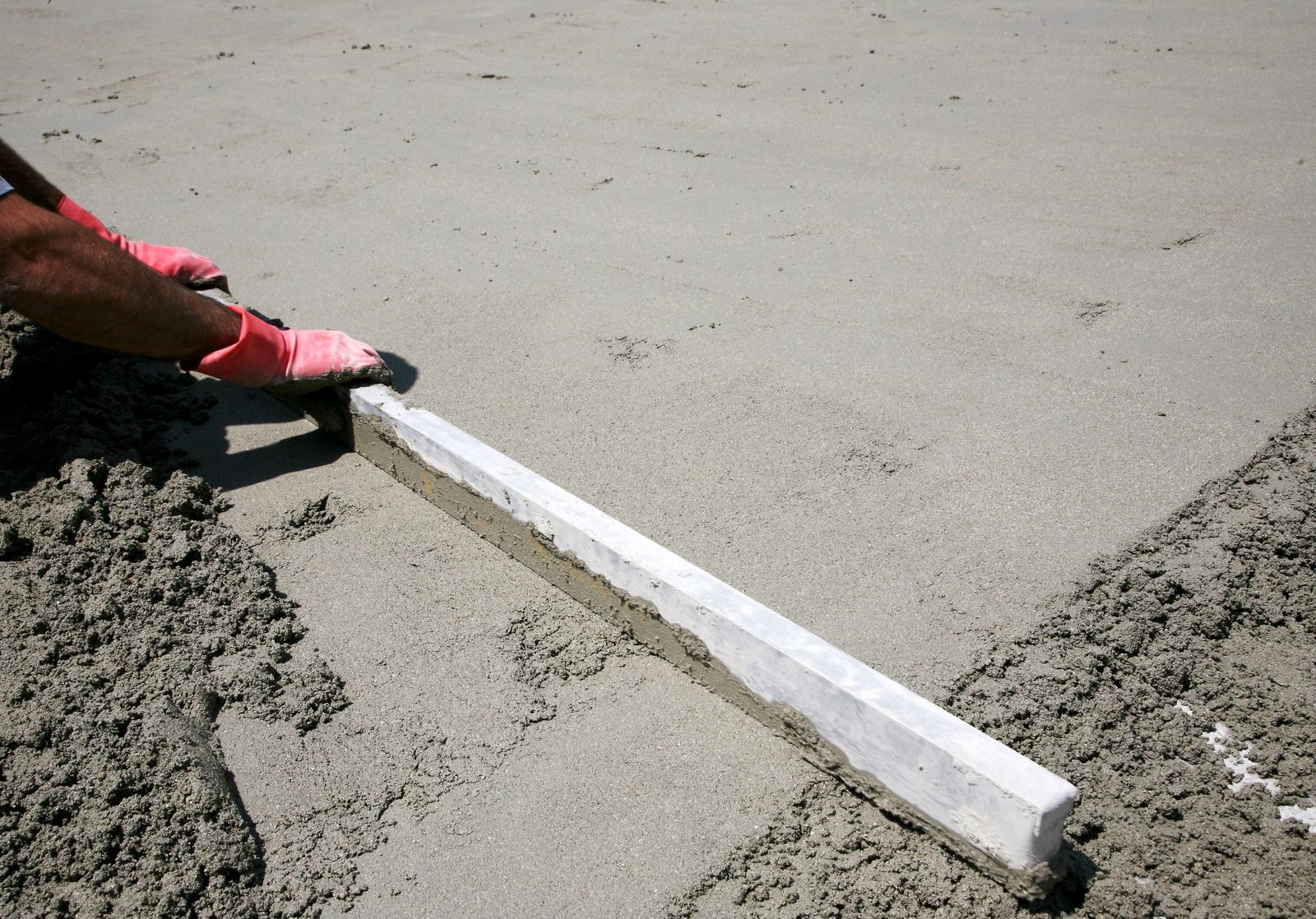 Person wearing pink gloves using a screed to level wet concrete.