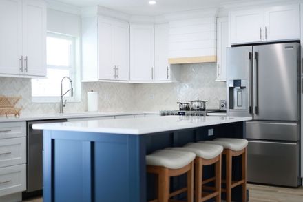 A modern kitchen featuring white cabinets, a navy blue island with bar stools, stainless steel appliances, and tile backsplash.