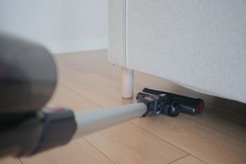 A vacuum cleaner nozzle cleans the light wooden floor underneath a white sofa.