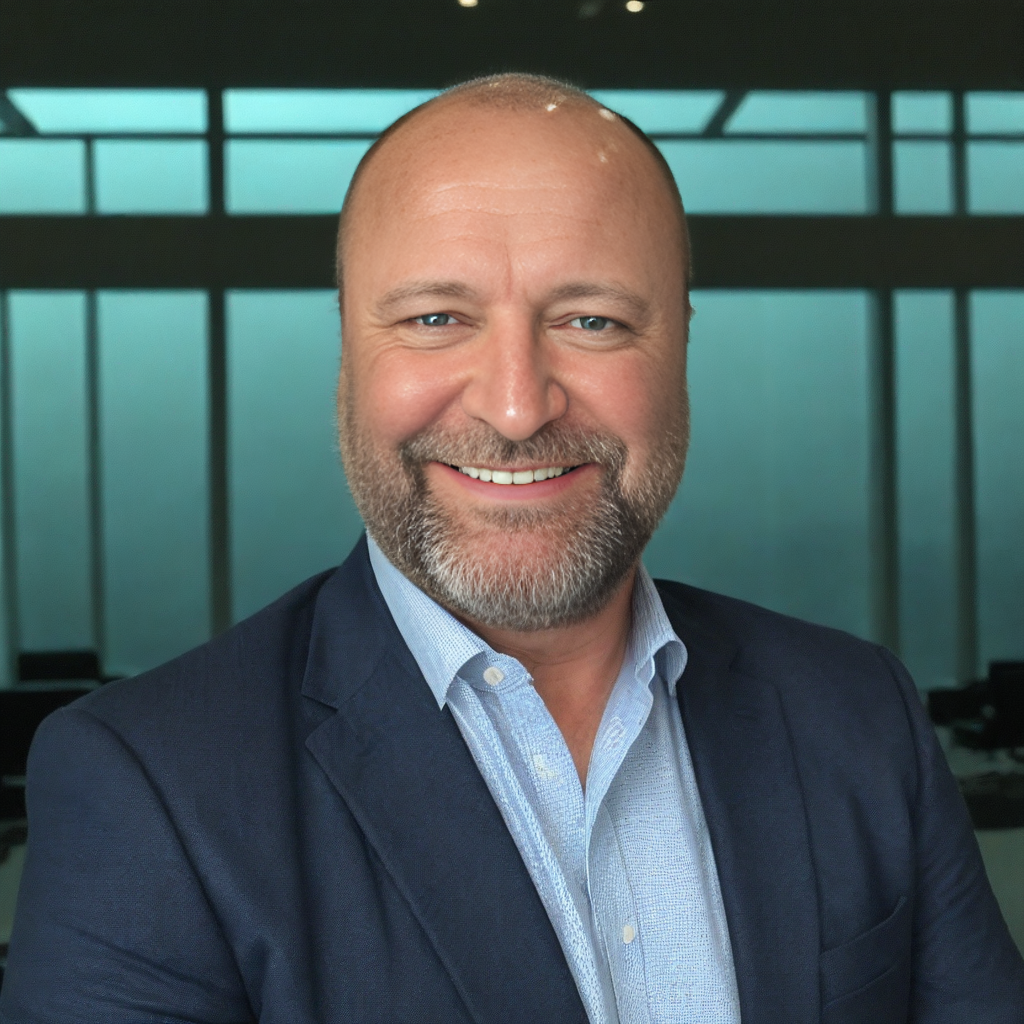 Man in a blue suit, white shirt, hands in pockets, standing in front of a modern home, green lawn.