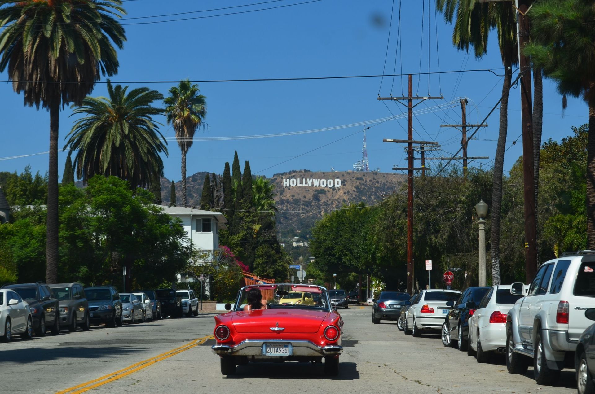 Red convertible driving down a palm-lined street with the Hollywood sign in the distance.