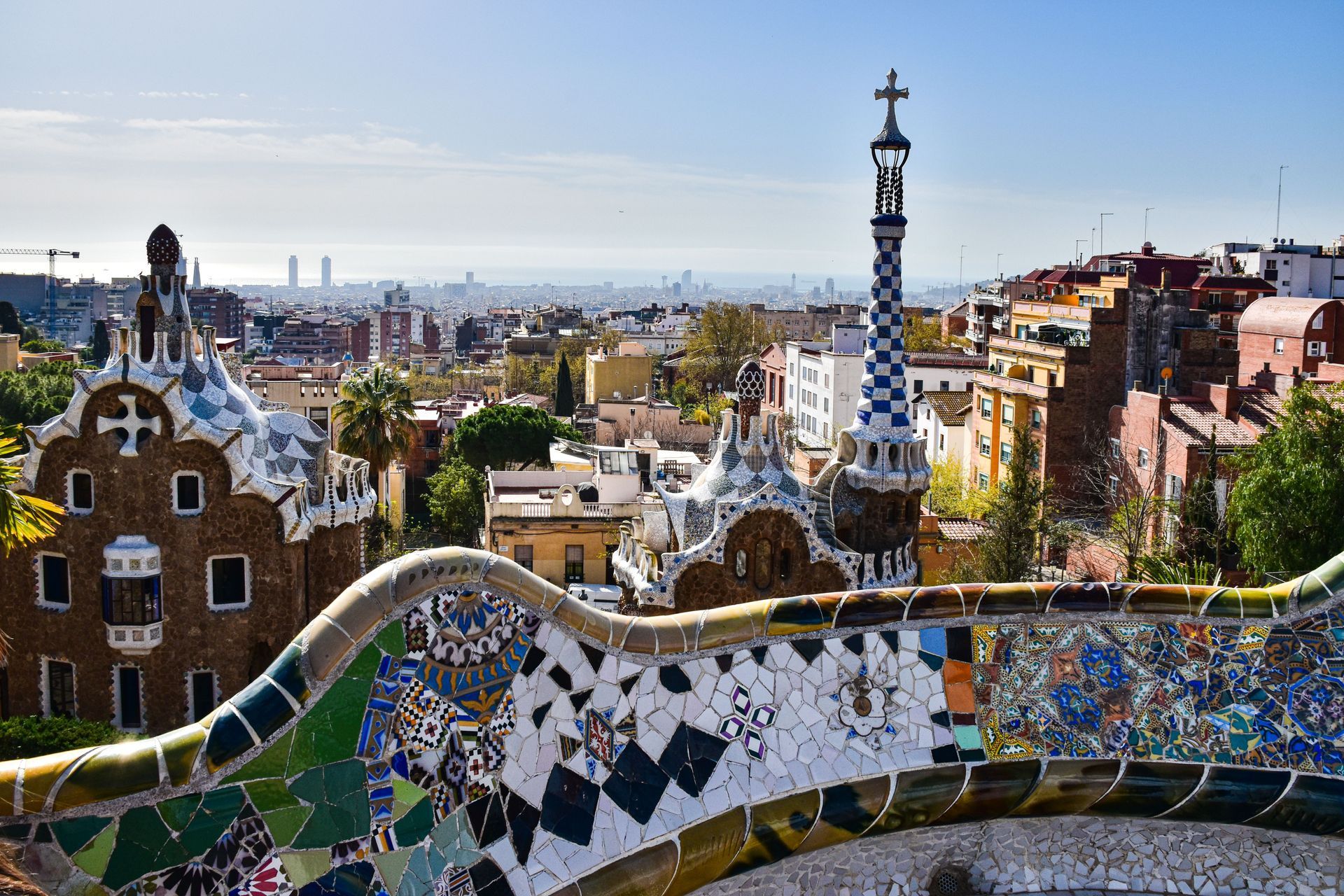 Colorful mosaic terrace overlooking a city skyline in Parc Güell, Barcelona, with a distant tower and bright blue sky