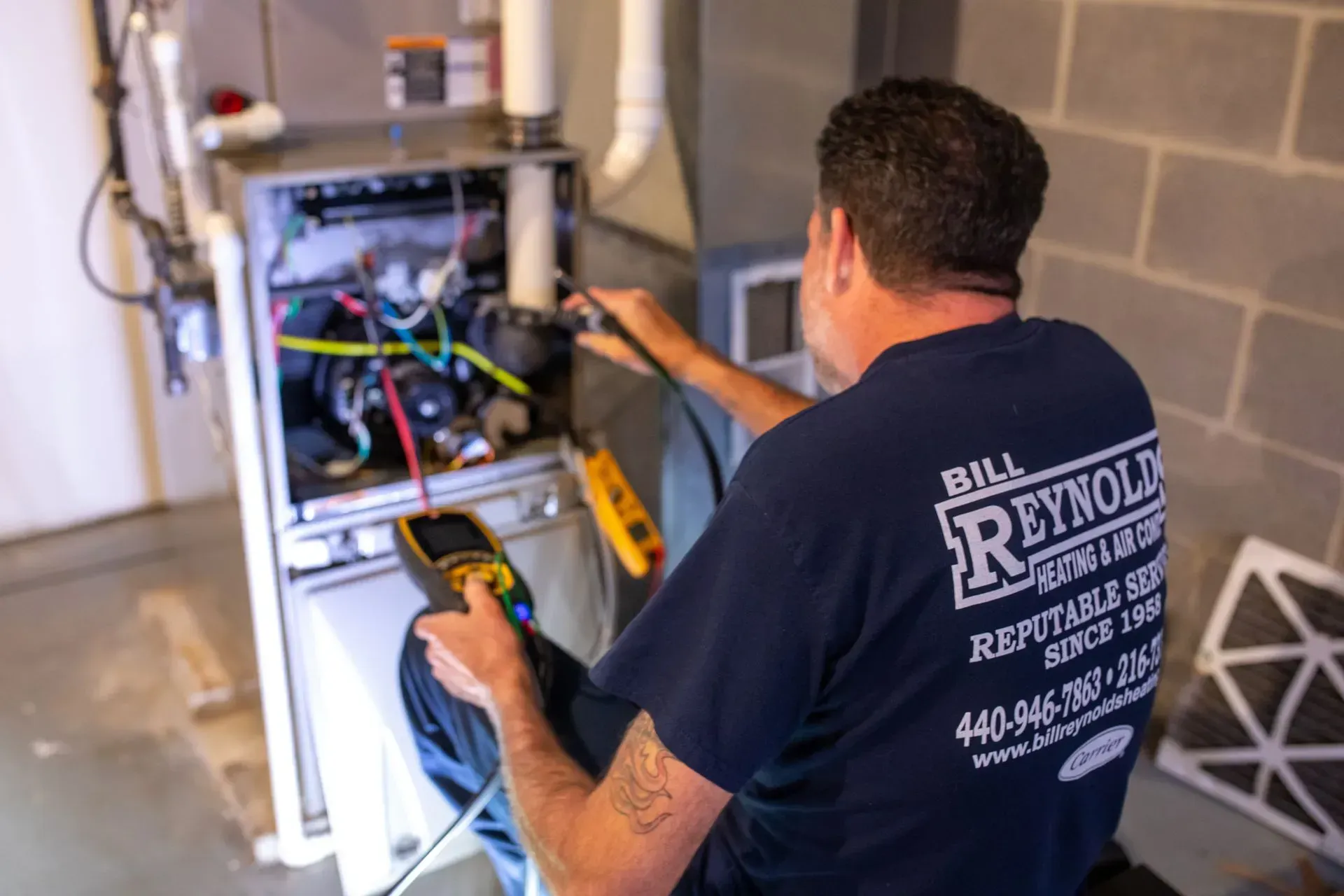 HVAC technician tests furnace with a multimeter. Grey-haired man in a blue shirt, inside.