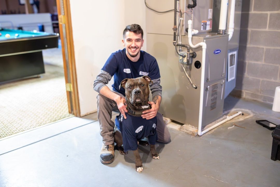 Man kneels with a dog in a basement. They wear matching blue work shirts. The man smiles near a furnace.