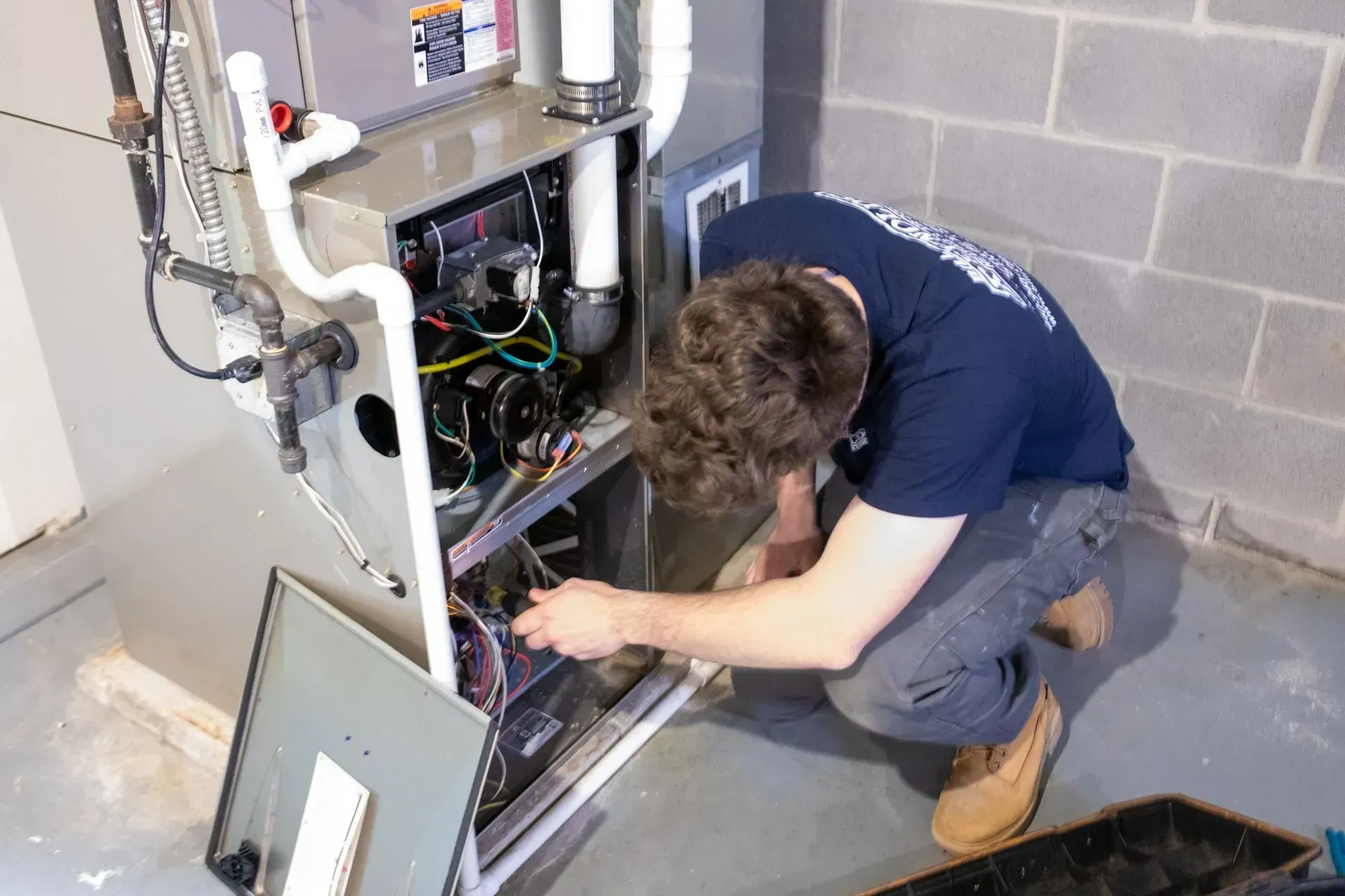 A man in blue shirt repairs a furnace in a basement. The furnace is gray.