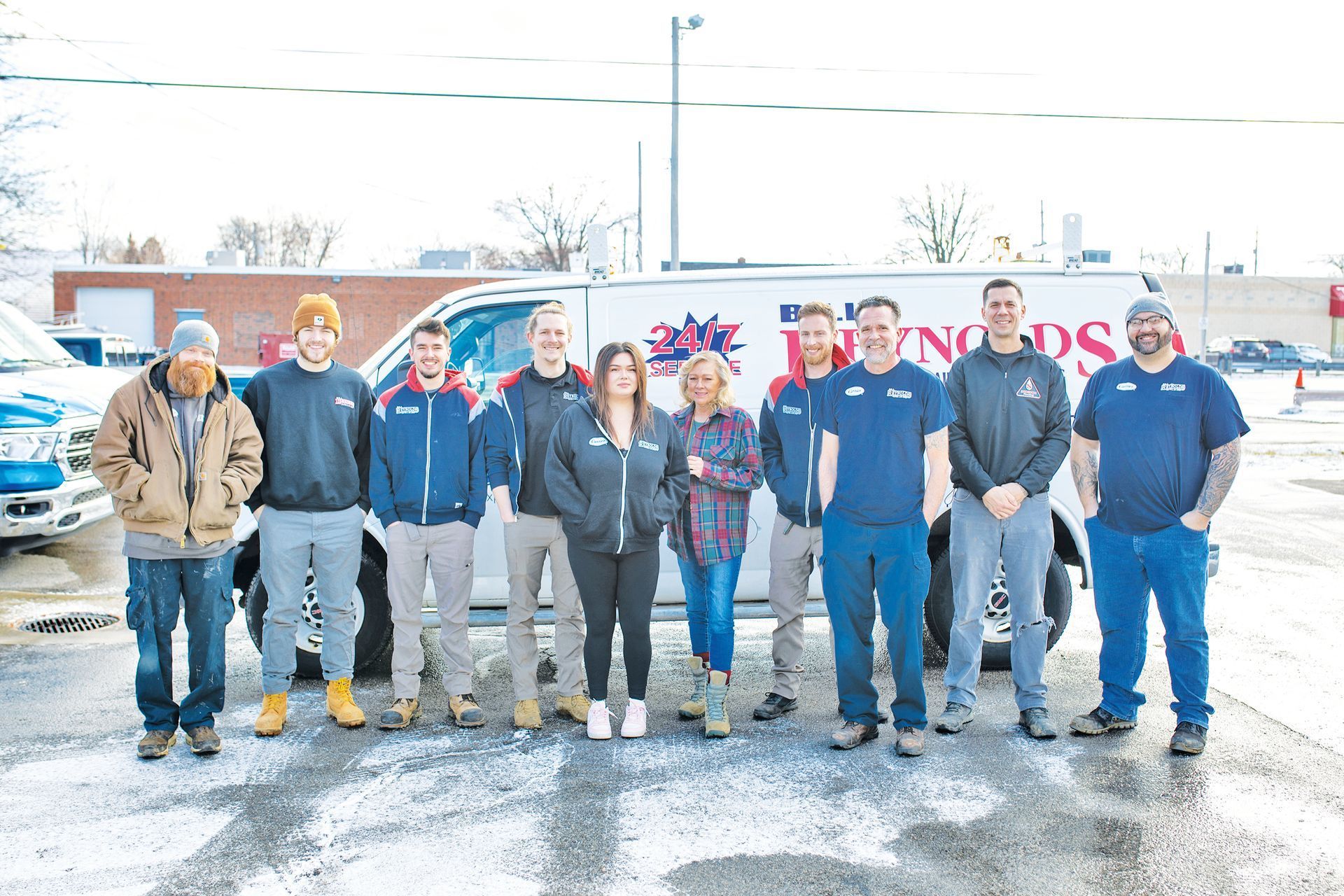 Team of 11 standing in front of Reynolds Heating van; some wear work uniforms, outdoors on a winter day.