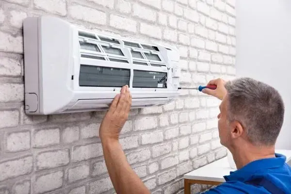 Man in blue shirt fixing a white air conditioner unit on a brick wall, using a screwdriver.