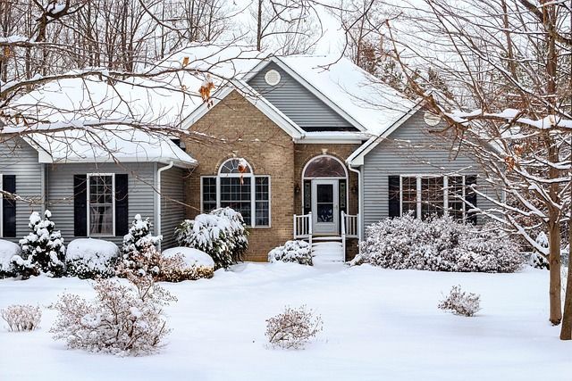 House covered in snow, surrounded by trees and bushes. Winter scene with gray and tan siding.