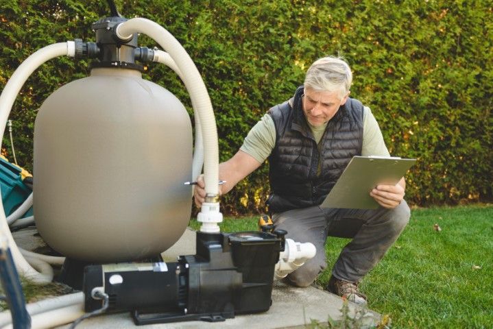 A person in a vest kneels outdoors, inspecting a pool pump and filter system while holding a clipboard.