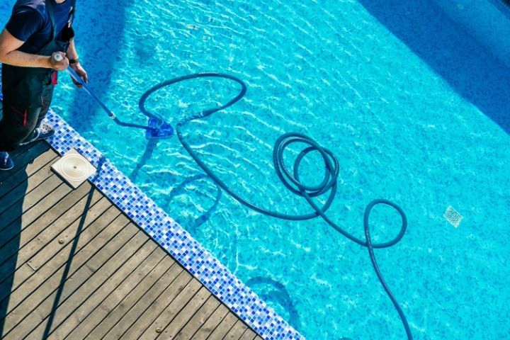 A person uses a long pole and hose to clean a vibrant blue tiled swimming pool.