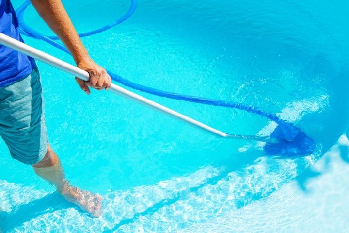 A person in shorts cleans a blue swimming pool with a long-handled brush and hose.