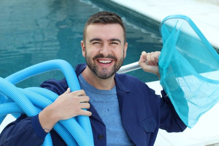A smiling pool technician holds a blue vacuum hose and a skimmer net beside a swimming pool.