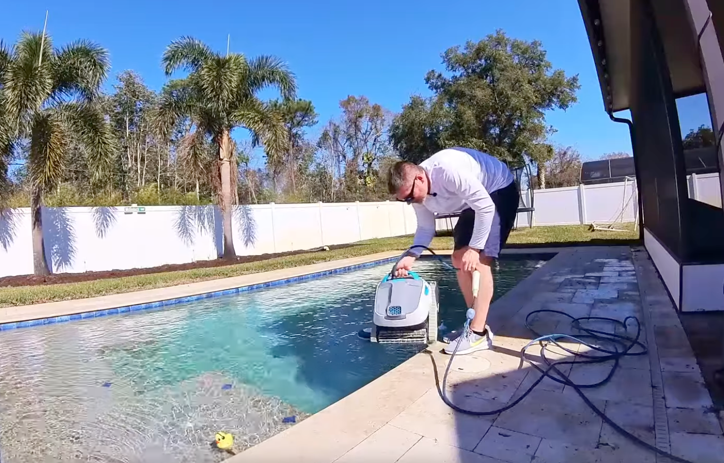 A person in a white long-sleeve shirt operates a robotic pool cleaner at the edge of a backyard swimming pool.