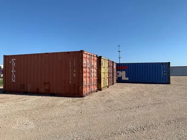 Stacked shipping containers in a sunny gravel lot, including a red container in front and a blue one behind.