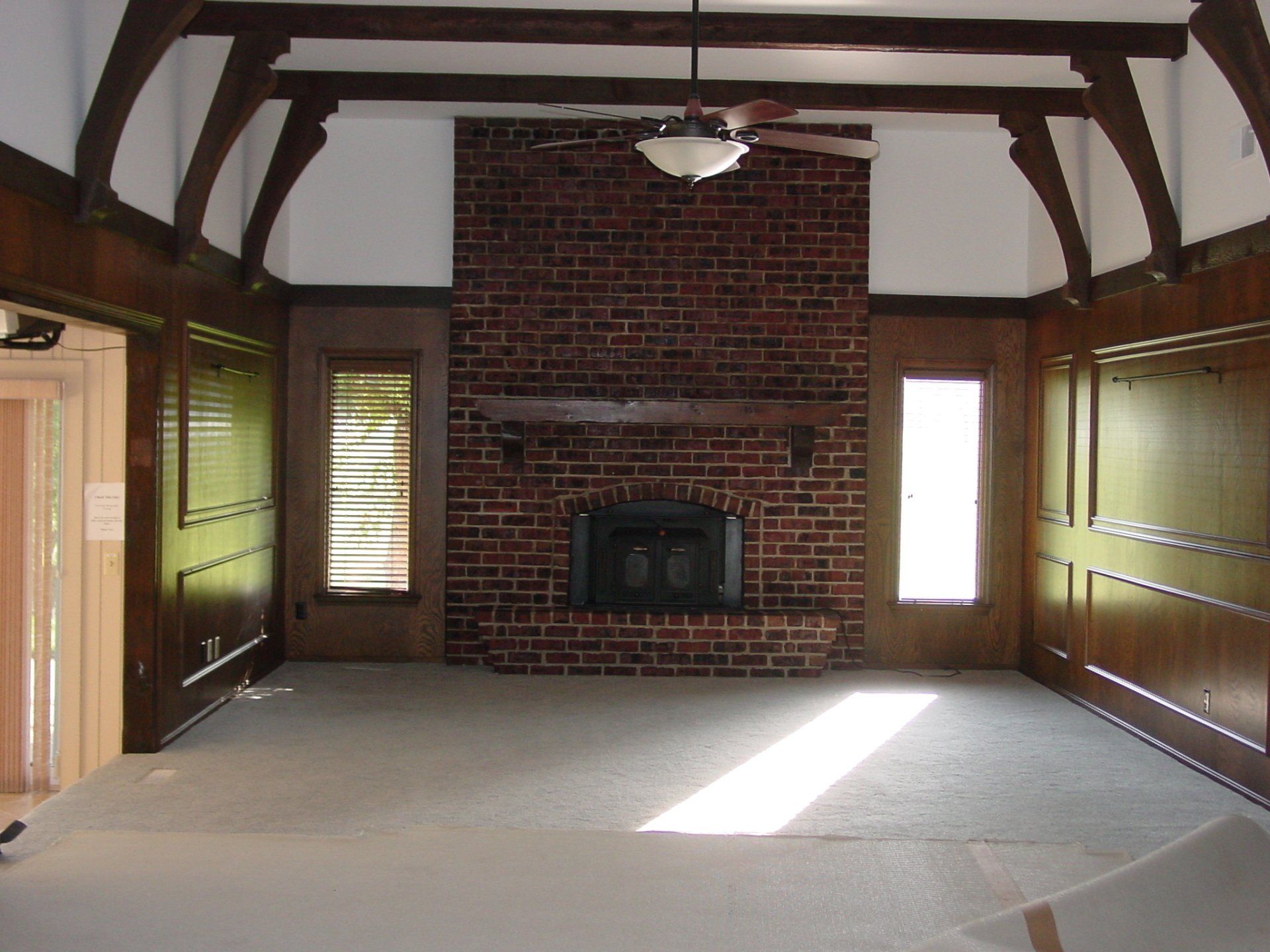 Empty living room with brick fireplace, dark wood paneling, and a high ceiling with exposed beams.