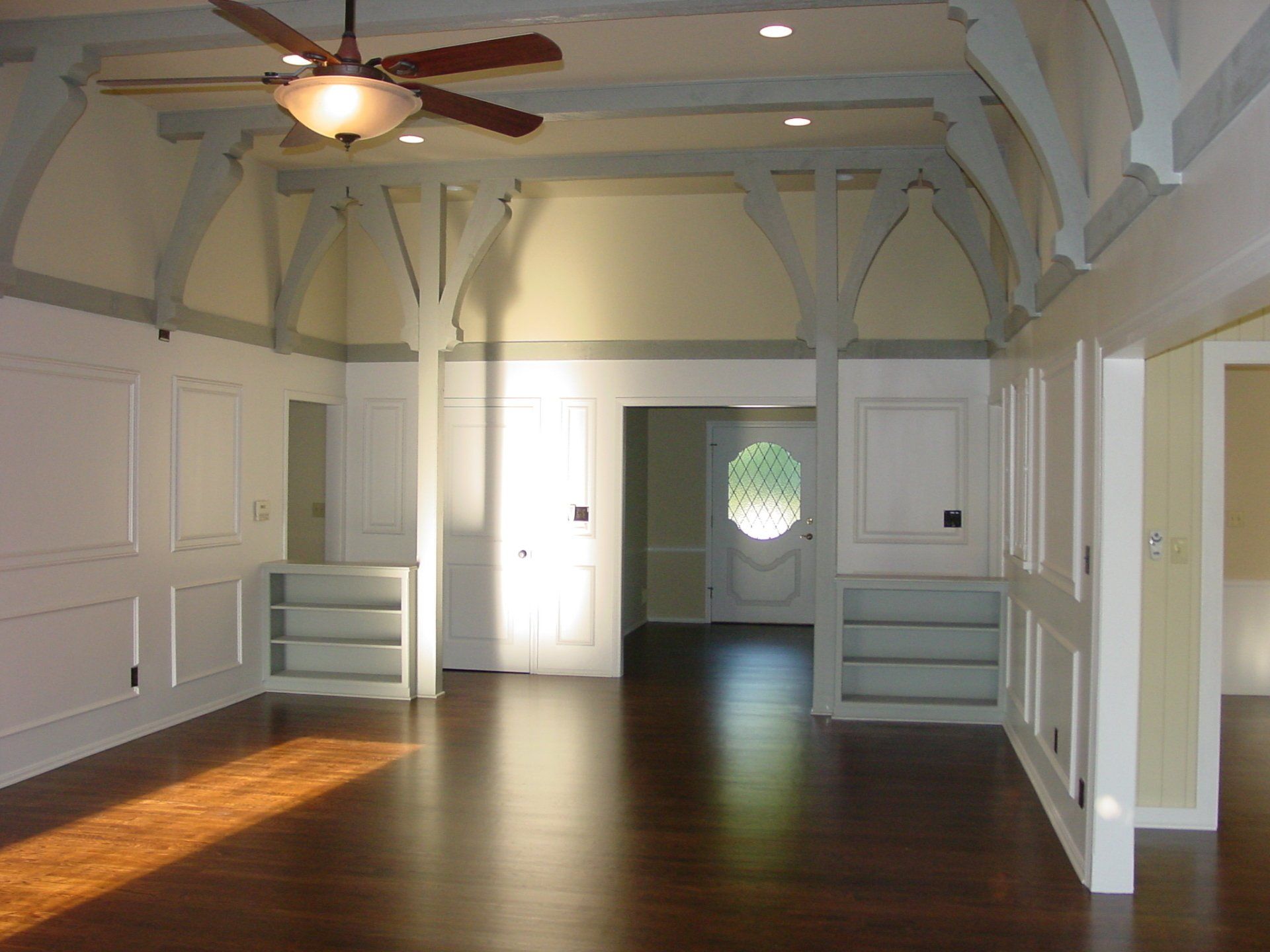 Spacious room with dark wood floors, arched supports, and white wainscoting. Light streams through a doorway.