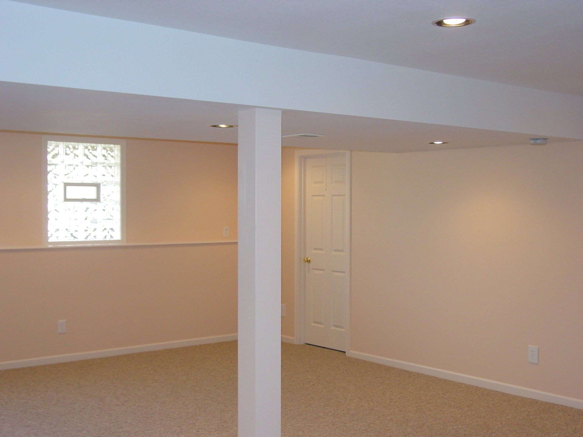 Empty basement with tan carpet, a window, white support beam, and a closed door.