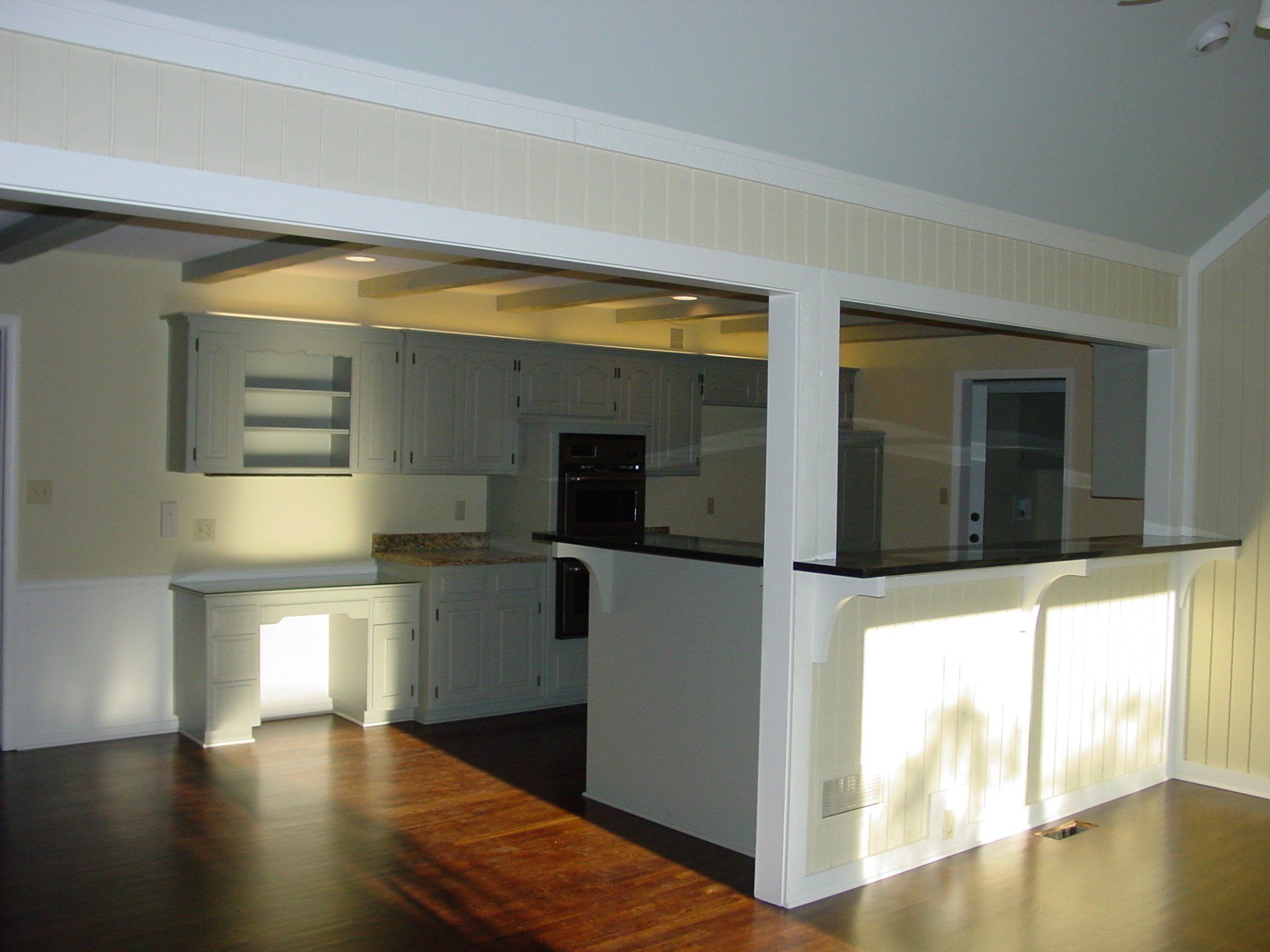 Interior view of a kitchen, open to a living area, with white cabinets, dark floors, and a bar.