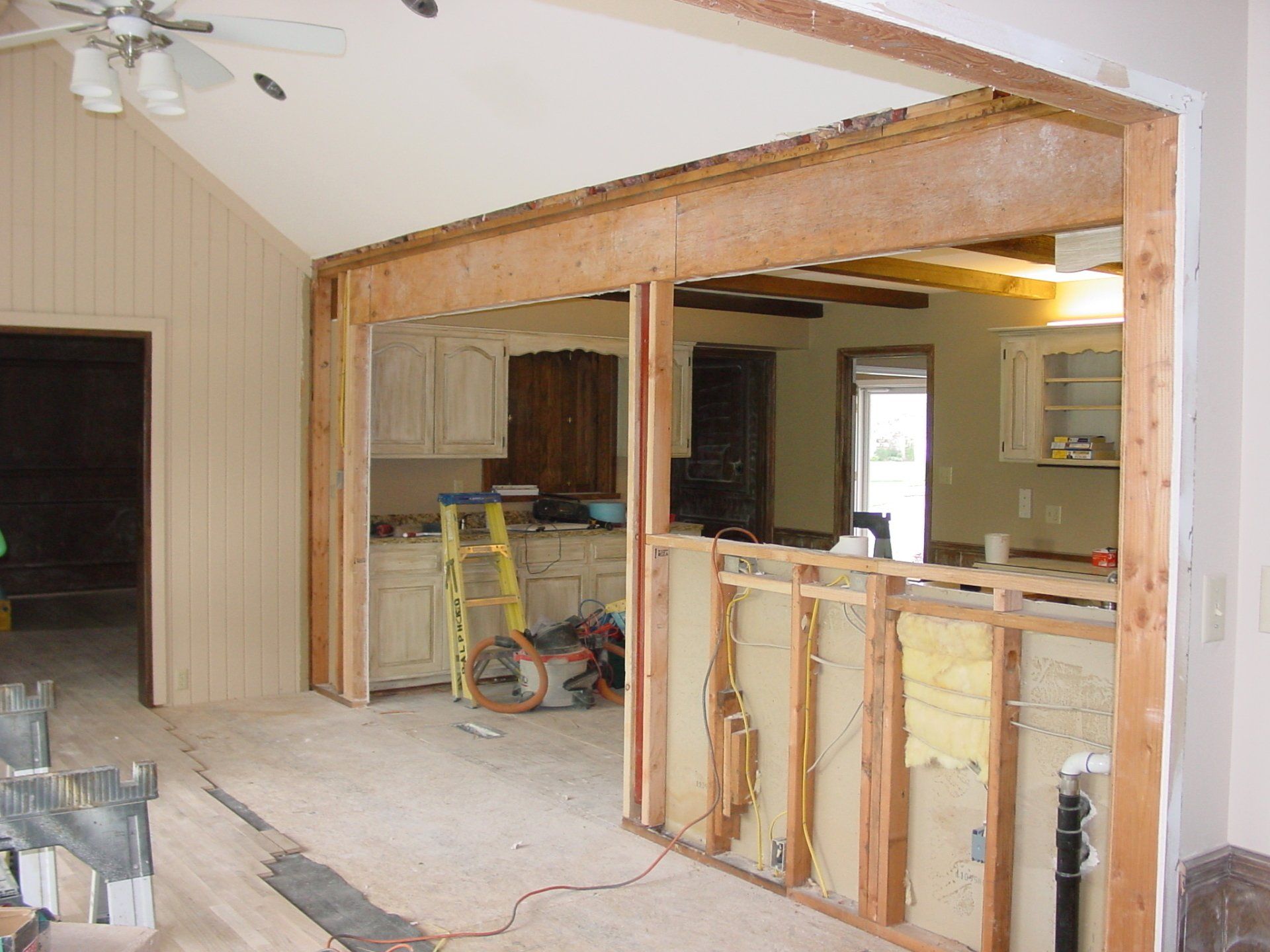 Interior renovation showing partial wall removal; wooden framing, exposed electrical, kitchen in background.