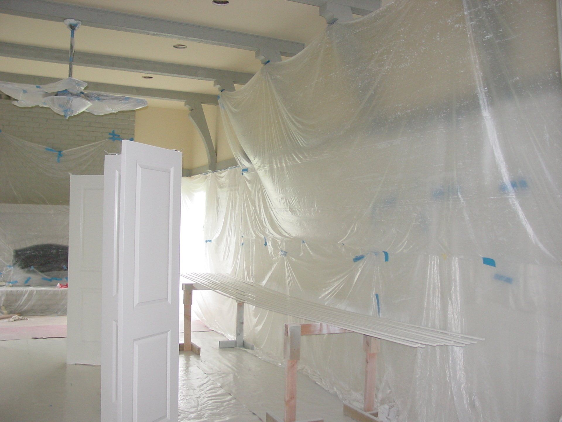 Interior room being painted, plastic sheeting covers walls, white door ajar, drying rack in front.