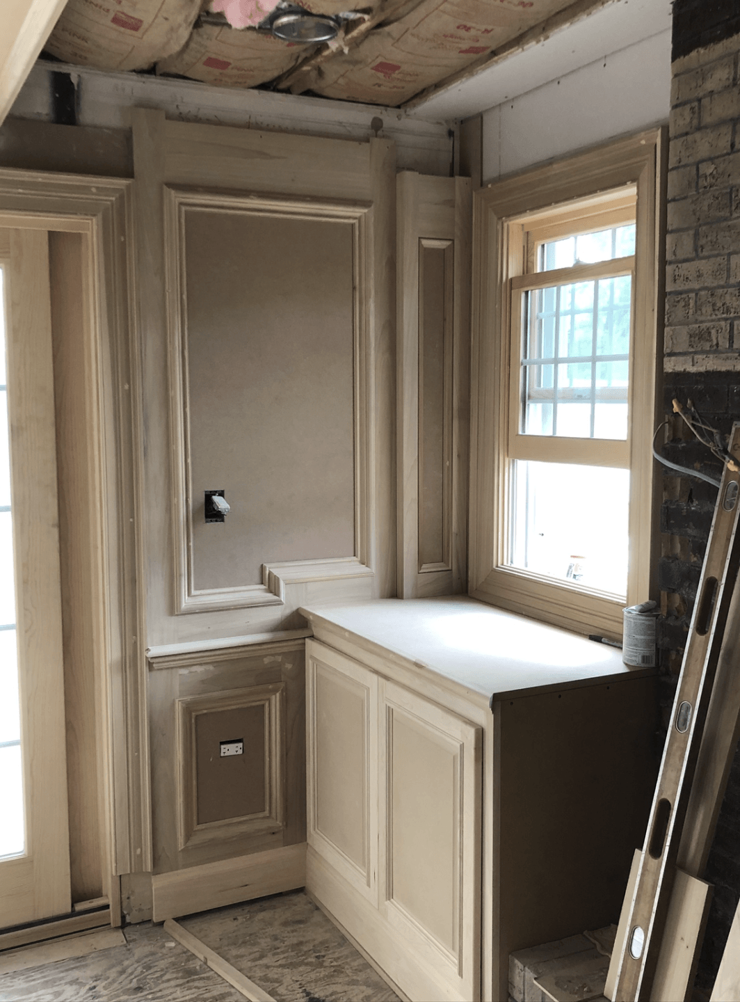 Interior under construction with wood trim, built-in cabinet, corner window, and bare brick wall.