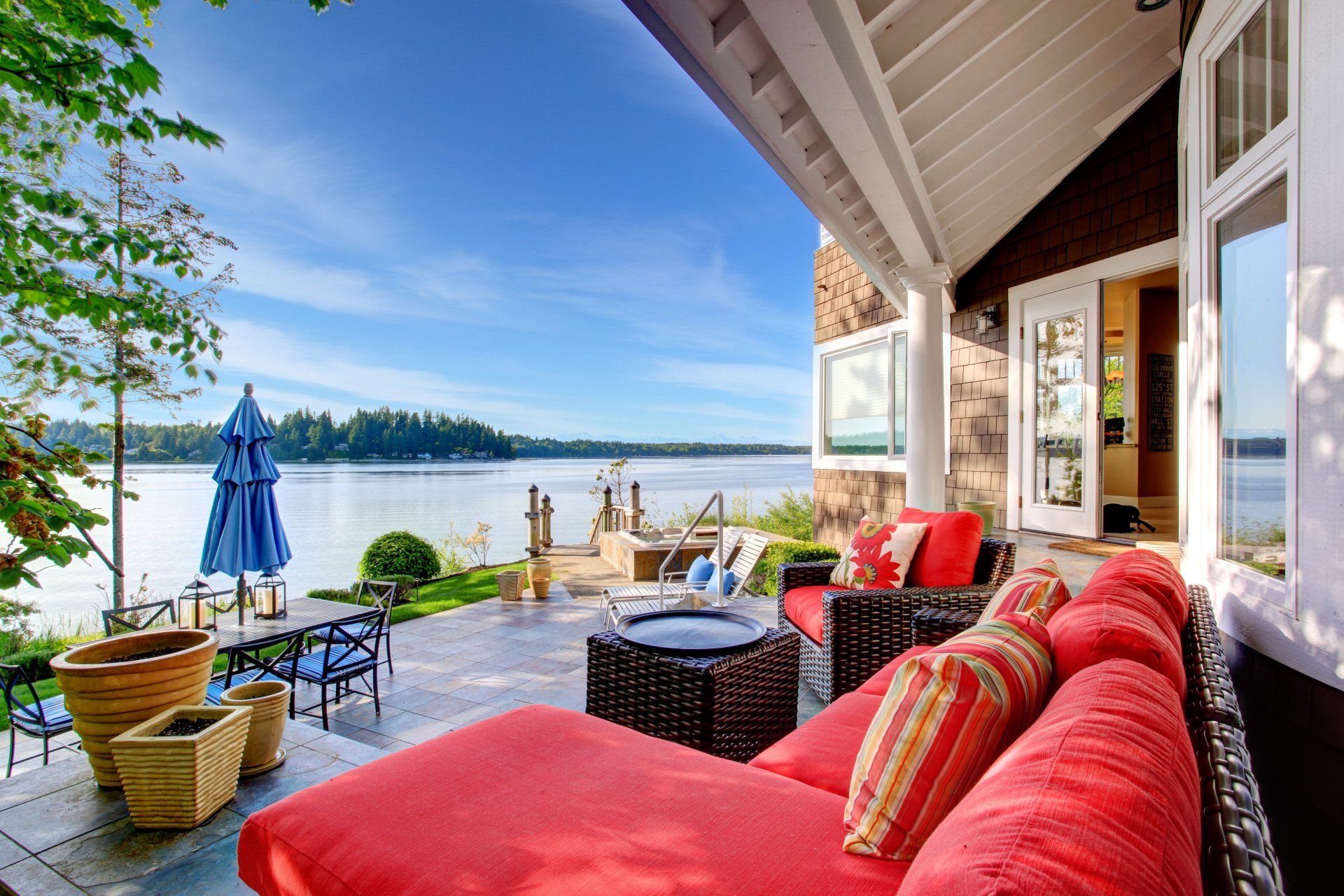 Patio with red couch, view of lake, blue umbrella, table, and plants. Sunny day.
