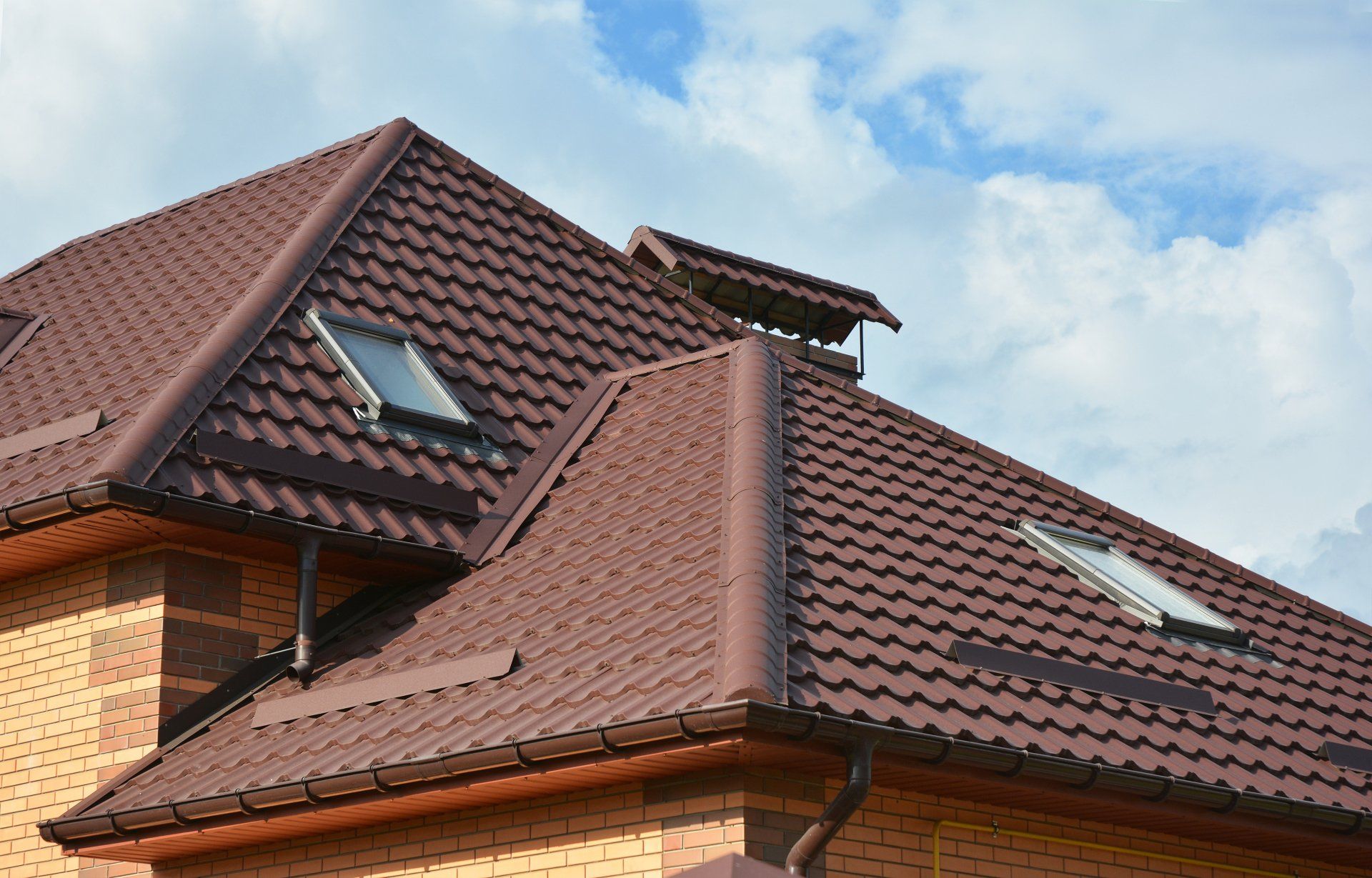 Brown metal roof on a brick building, with skylights and a chimney against a cloudy sky.
