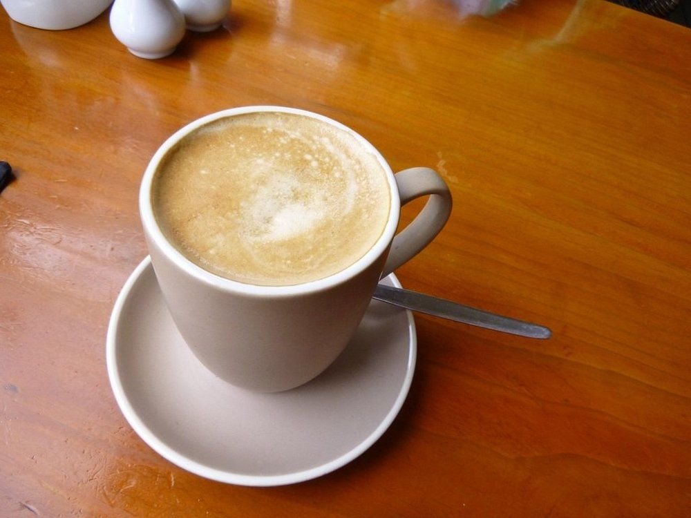 A Cappuccino in A Beige Mug and Saucer on A Polished Wooden Table with A Spoon — Kape Centro In Childers, QLD