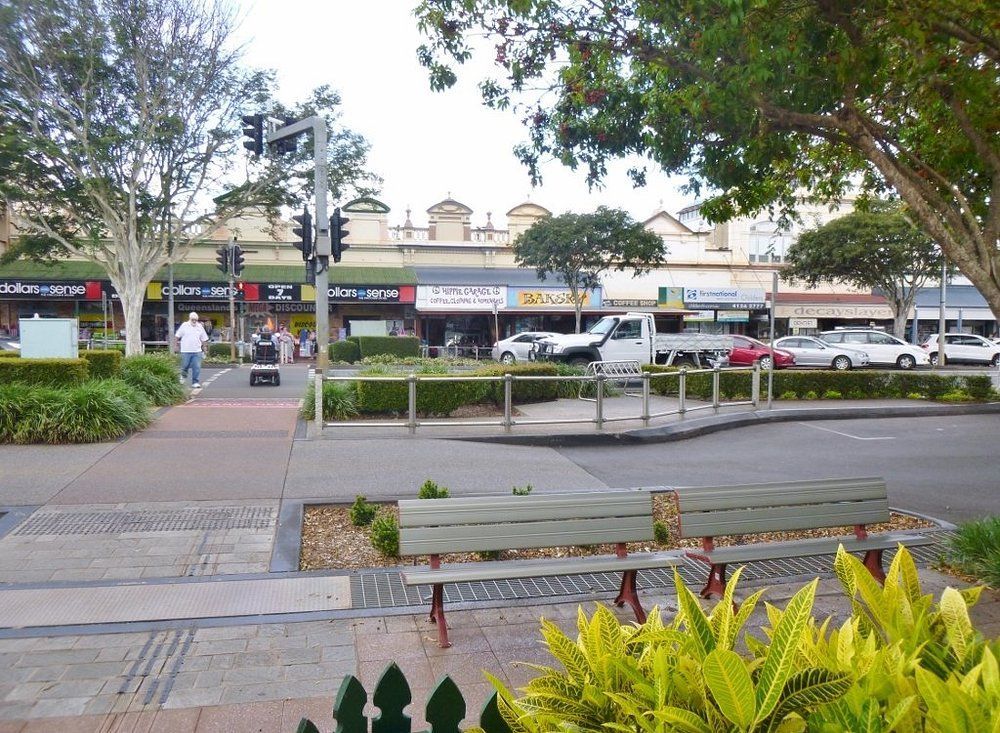 Street Scene with Shops, Pedestrians, Trees, and A Bench — Kape Centro In Childers, QLD