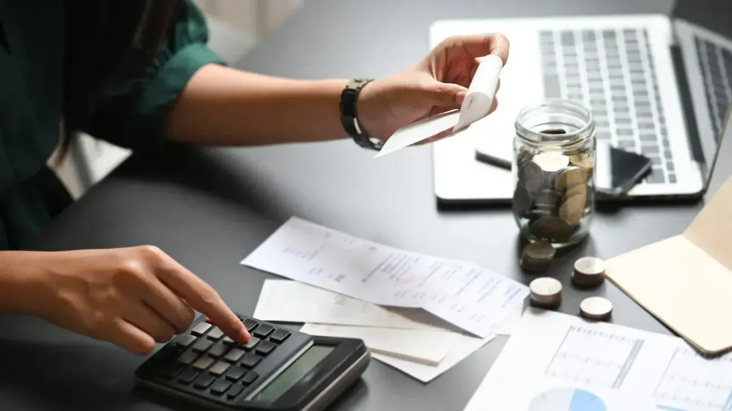 A person calculates expenses using a calculator, receipts, and a jar of coins on a desk