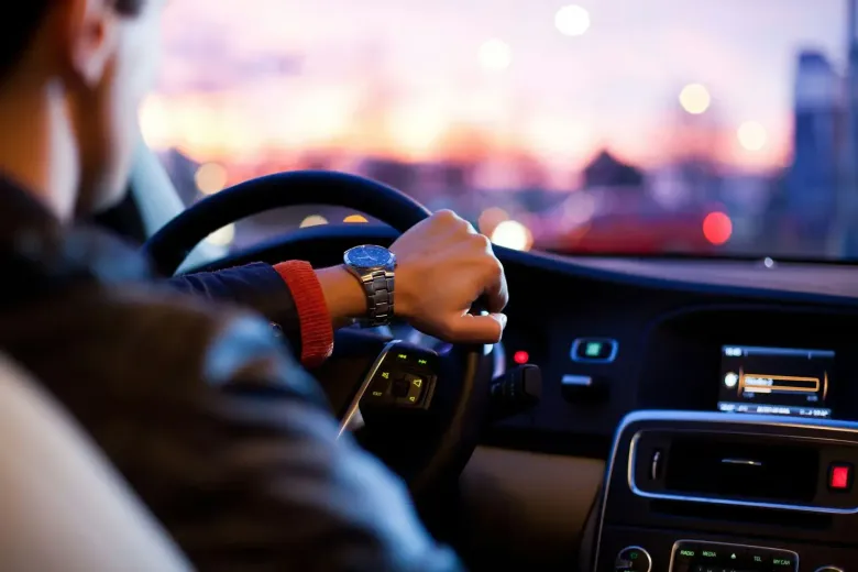 A driver's hands on a steering wheel inside a car at sunset, with a blurred cityscape