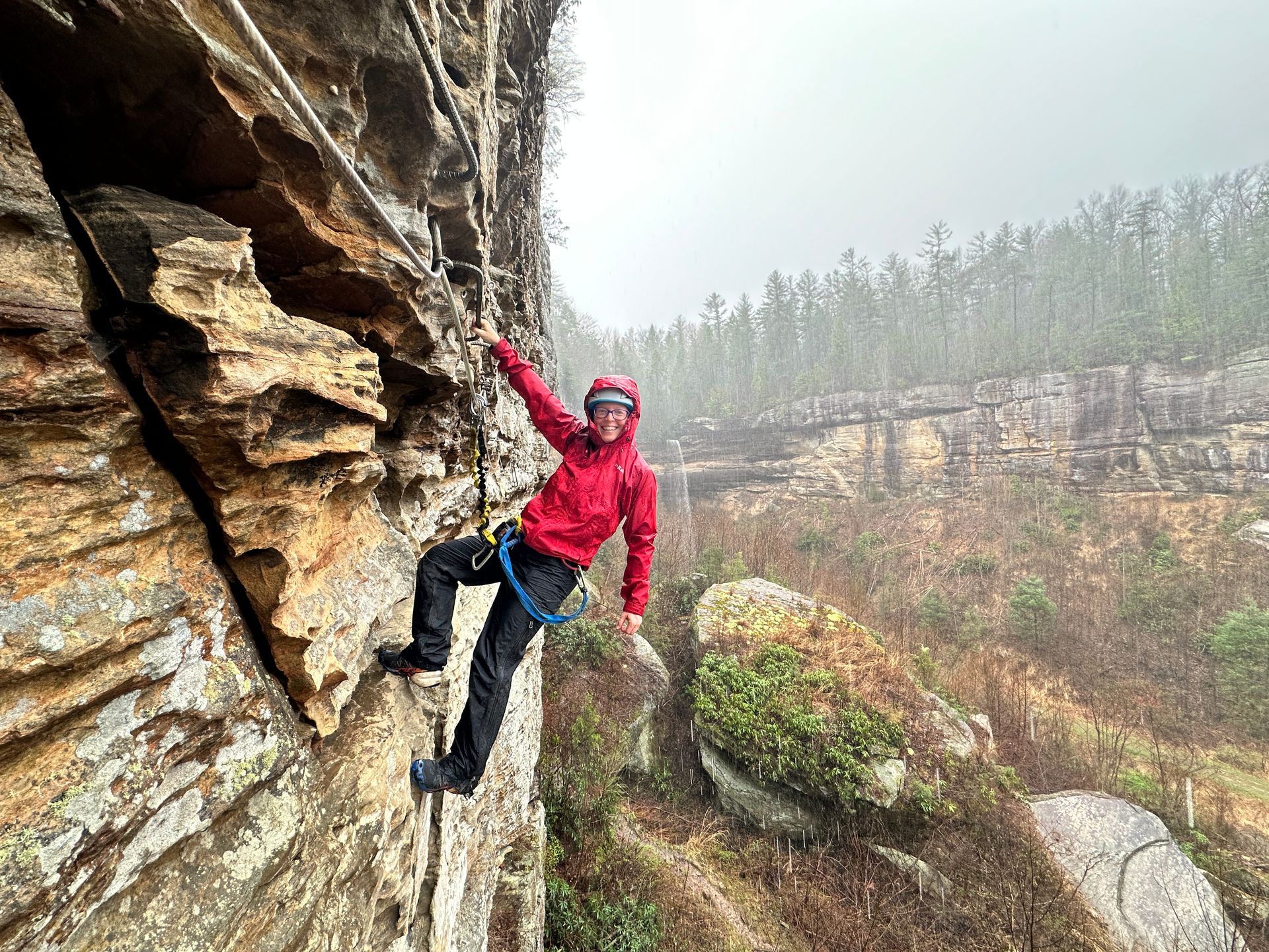 Woman climbing the Red River Gorge Via Ferrata in the rain, clipped into the safety cable as water f