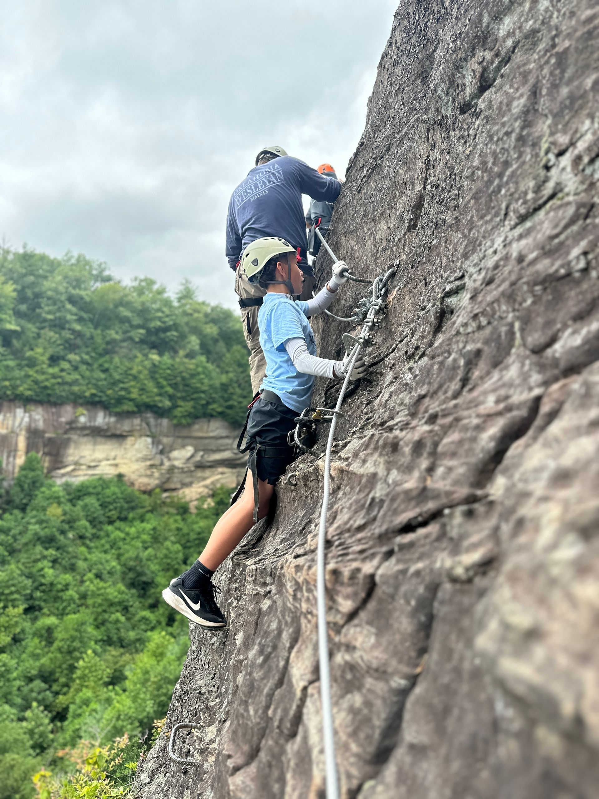 Father and son climb side by side on the Red River Gorge Via Ferrata, securely clipped into safety cables as they ascend iron rungs on a sandstone cliff.