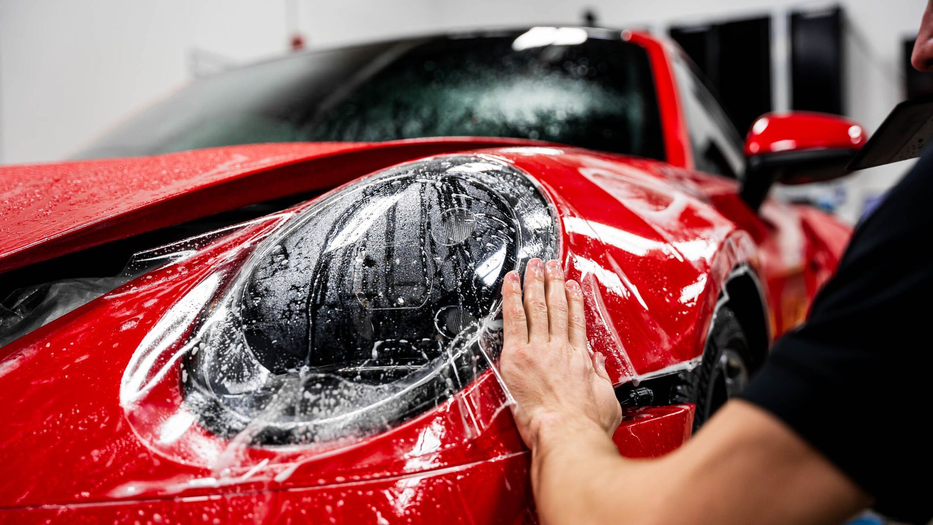 A man is applying protective film to the headlight of a red car.