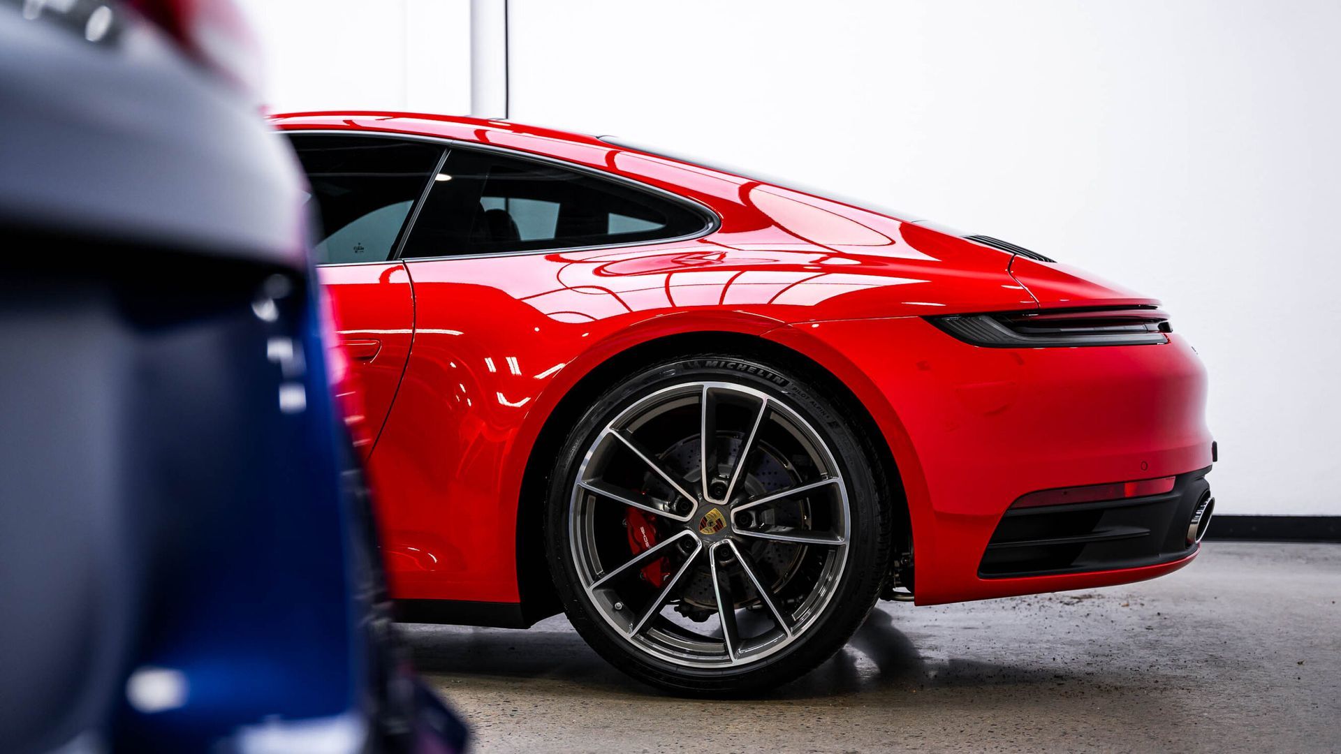 A red porsche 911 is parked next to a blue car in a garage.