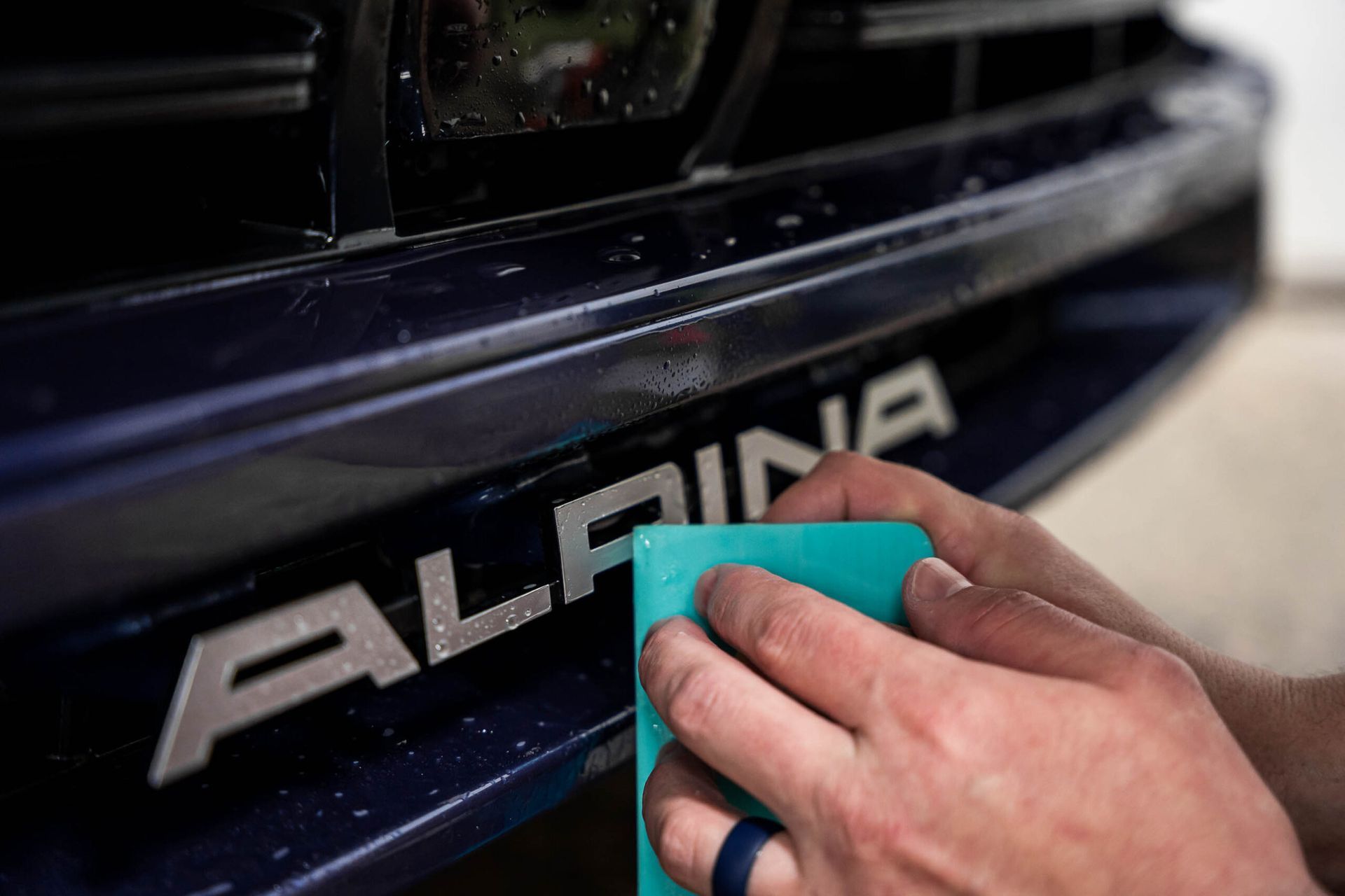 A person is cleaning the front of a blue alpina car with a sponge.