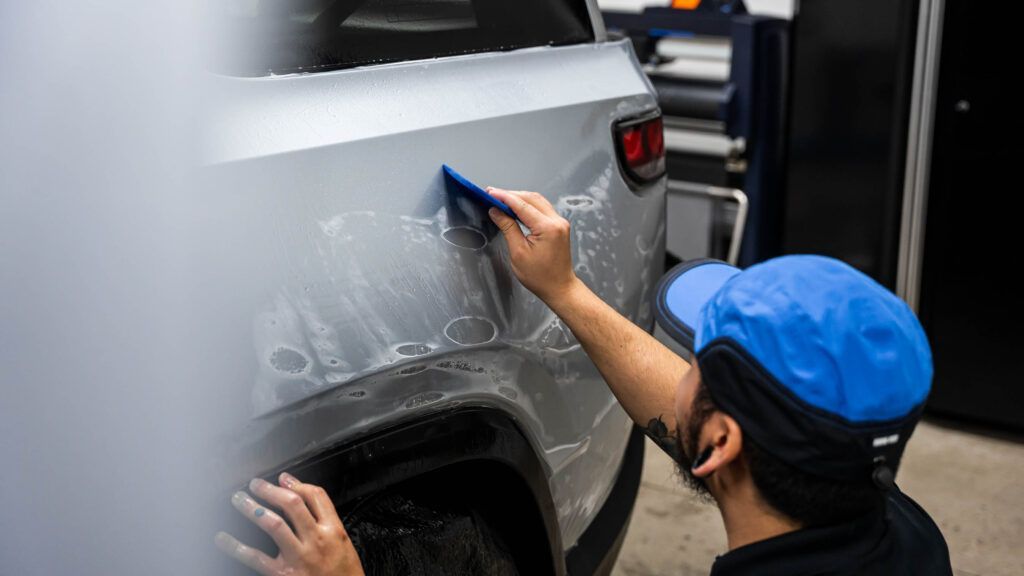 A man in a blue hat is applying a protective film to the side of a car.