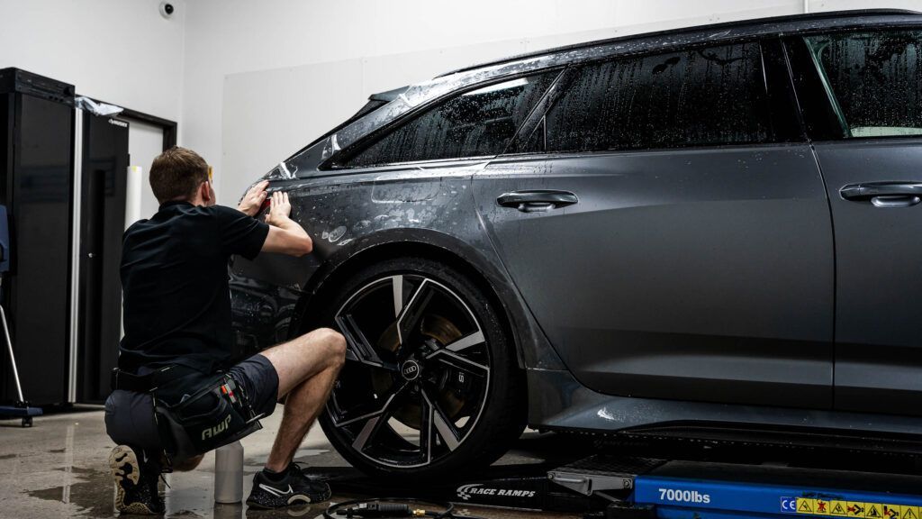 A man is kneeling down next to a car on a lift in a garage.