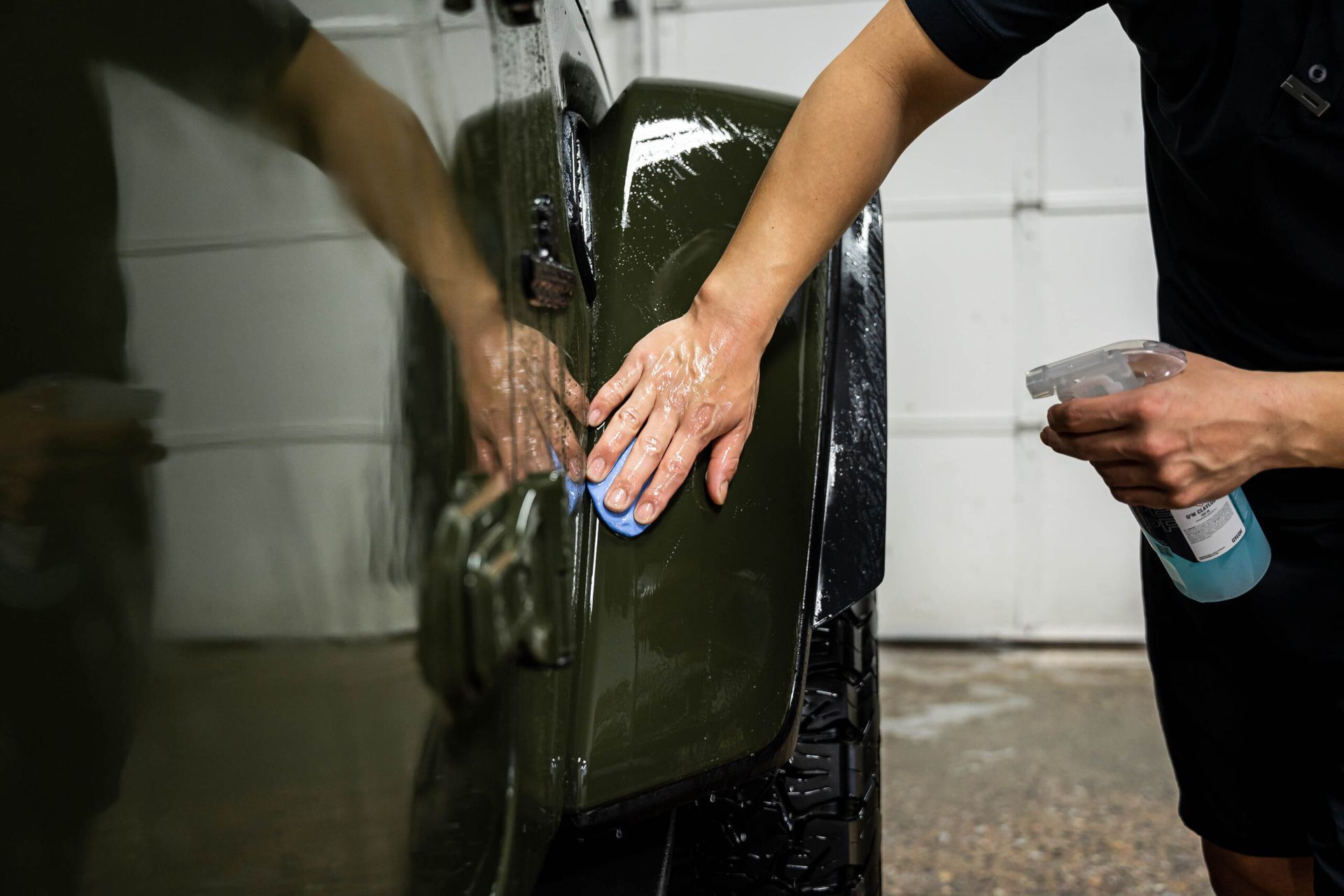 A person is washing a car in a garage while holding a spray bottle.