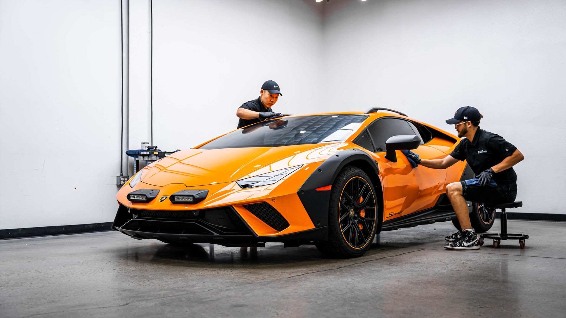 Two men are working on an orange sports car in a garage.