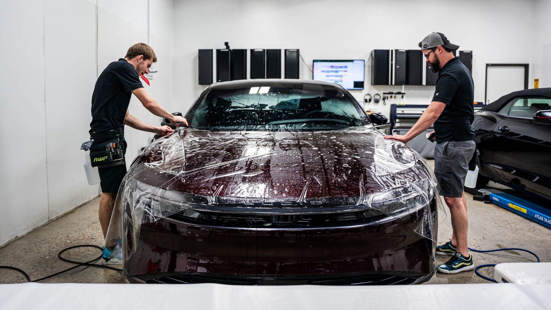 Two men are wrapping a car with plastic wrap in a garage.