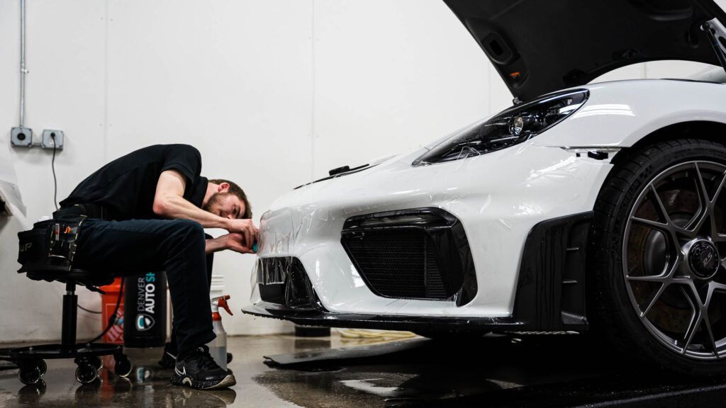 A man is sitting on a stool next to a white sports car with the hood open.