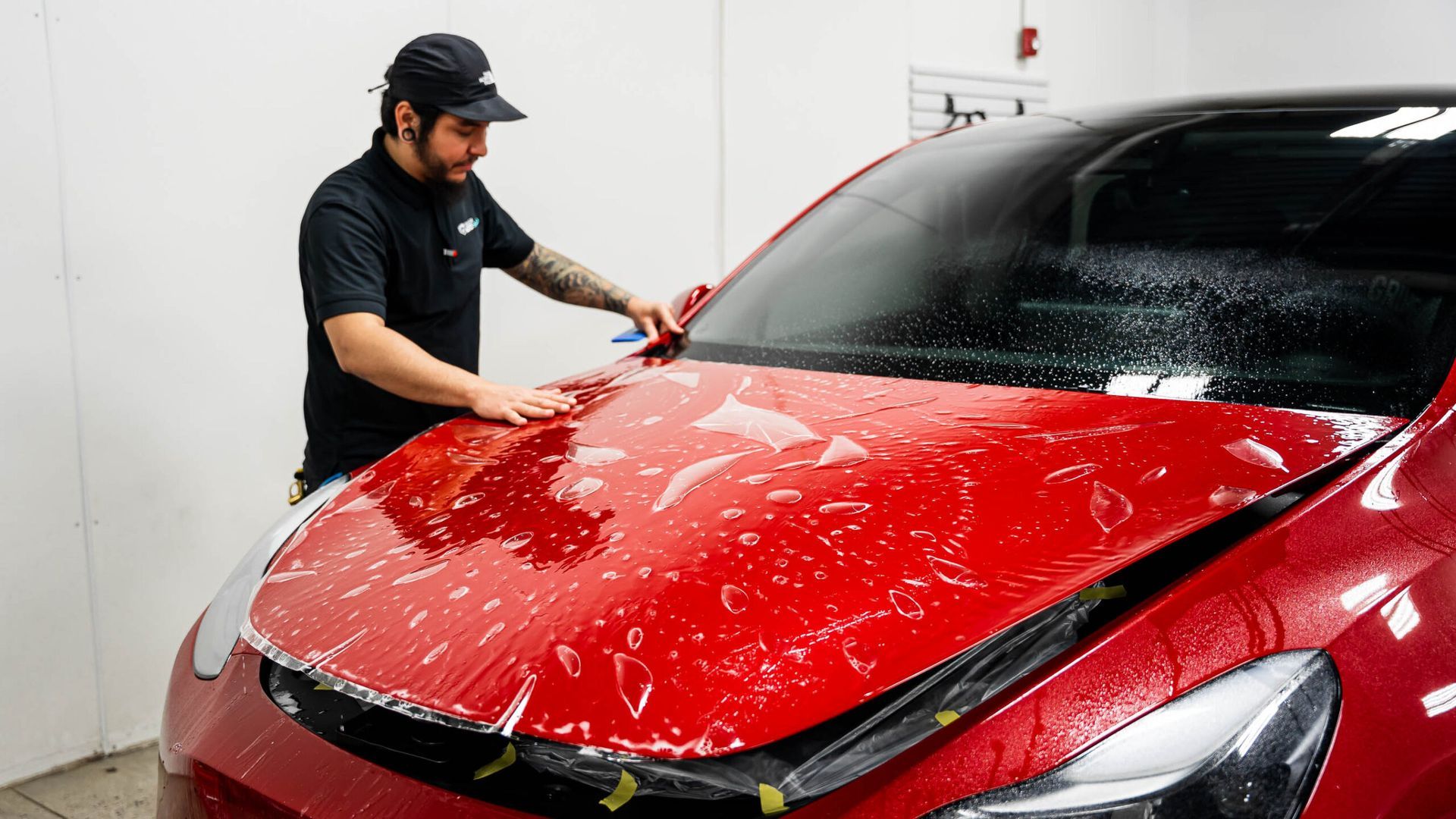 A man is applying a protective film to the hood of a red car.