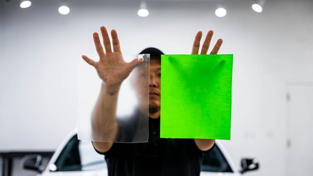 A man is holding a green and a clear piece of plastic in front of a car.